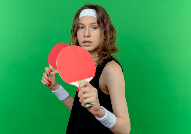 Chica joven fitness en ropa deportiva negra con diadema sosteniendo dos raquetas de tenis de mesa con cara seria de pie sobre la pared verde