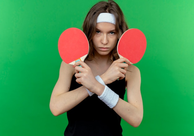 Chica joven fitness en ropa deportiva negra con diadema sosteniendo dos raquetas de tenis de mesa disgustado cruzando las manos de pie sobre la pared verde