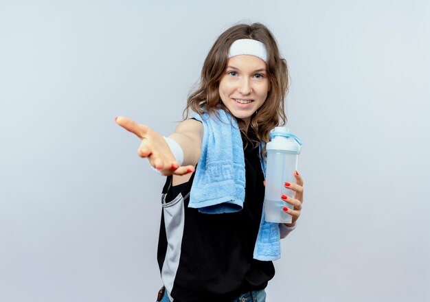 Chica joven fitness en ropa deportiva negra con diadema y toalla alrededor del cuello sosteniendo una botella de agua haciendo gesto de ven aquí con la mano sonriendo de pie sobre la pared blanca