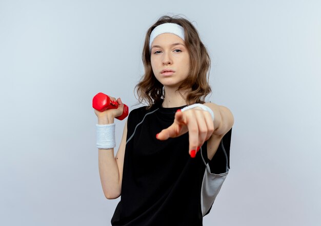 Chica joven fitness en ropa deportiva negra con diadema trabajando con mancuernas con cara seria apuntando con el dedo parado sobre la pared blanca
