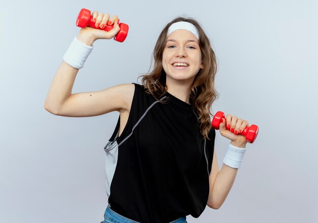 Chica joven fitness en ropa deportiva negra con diadema trabajando con pesas sonriendo de pie sobre la pared blanca