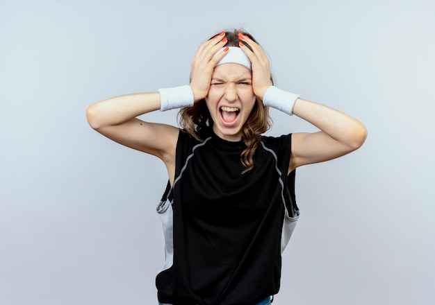 Foto gratuita chica joven fitness en ropa deportiva negra con diadema volviéndose salvaje sosteniendo su cabeza gritando de pie sobre la pared blanca