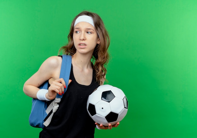 Chica joven fitness en ropa deportiva negra con mochila y diadema sosteniendo fútbol mirando a un lado confundido parado sobre pared verde