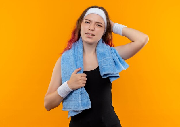 Chica joven fitness en ropa deportiva con una toalla en el cuello mirando confundida y muy ansiosa rascándose la cabeza de pie sobre la pared naranja