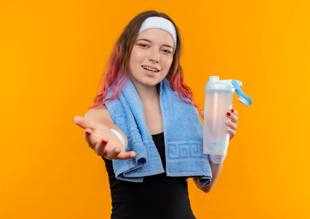 Chica joven fitness en ropa deportiva con una toalla en el cuello sosteniendo una botella con agua haciendo venir en gesto con la mano sonriendo alegremente de pie sobre la pared naranja