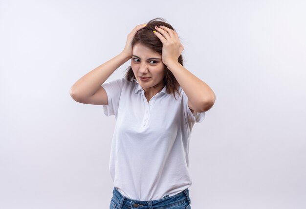 Chica joven frustrada con pelo corto vistiendo polo blanco tocando la cabeza muy enojado