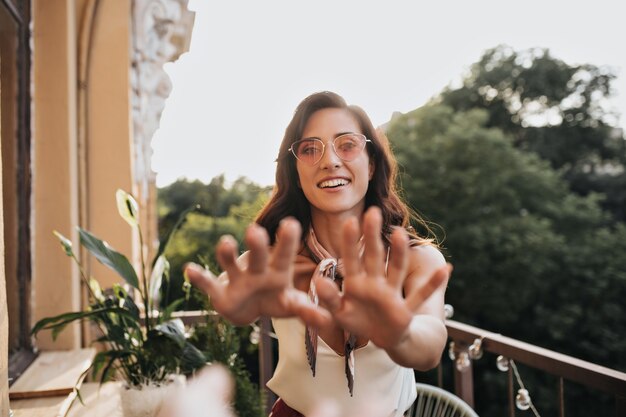 Chica joven con gafas de sol llega a la cámara. Hermosa mujer con cabello ondulado oscuro en blusa blanca está sonriendo y posando en la terraza.