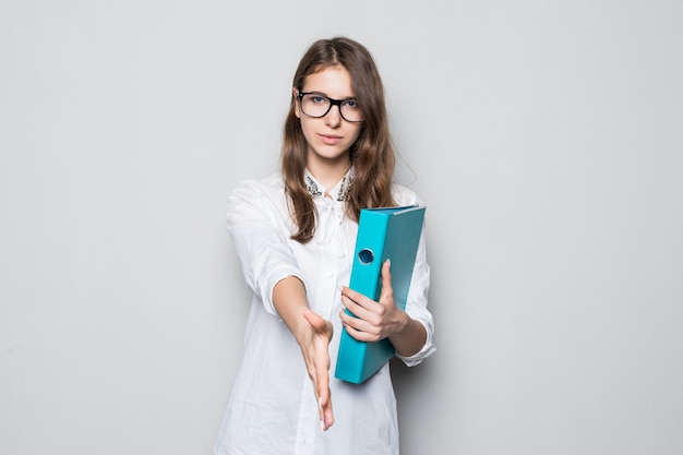 Chica joven con gafas vestidas con una estricta camiseta blanca de oficina se encuentra frente a una pared blanca con una carpeta azul para documentos en sus manos