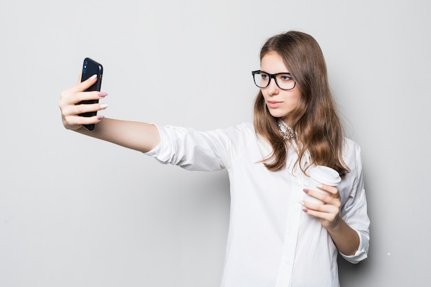 Chica joven con gafas vestidas con una estricta camiseta blanca de oficina se encuentra frente a una pared blanca y sostiene su teléfono en las manos