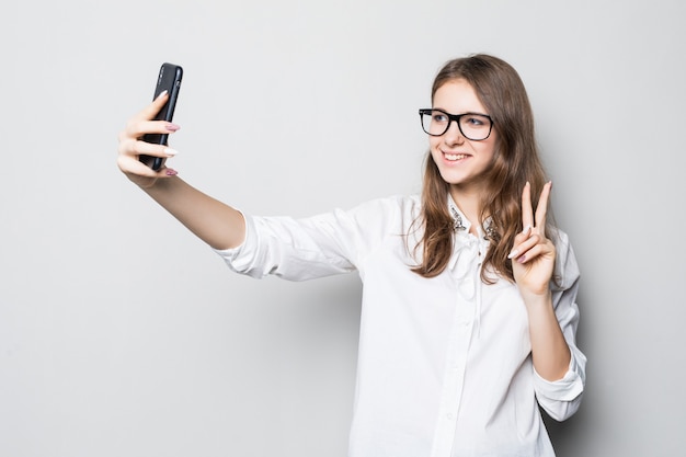 Chica joven con gafas vestidas con una estricta camiseta blanca de oficina se encuentra frente a una pared blanca y sostiene su teléfono en las manos