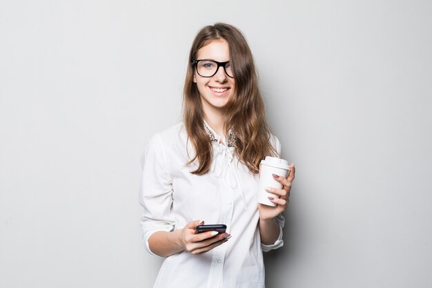 Chica joven con gafas vestidas con estricta camiseta blanca de oficina se encuentra frente a una pared blanca y sostiene su teléfono y una taza de café en las manos