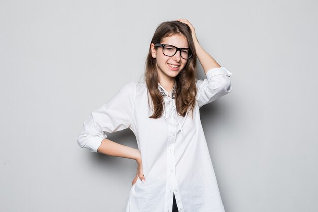 Chica joven con gafas vestidas con una estricta camiseta blanca de oficina se encuentra frente a la pared blanca