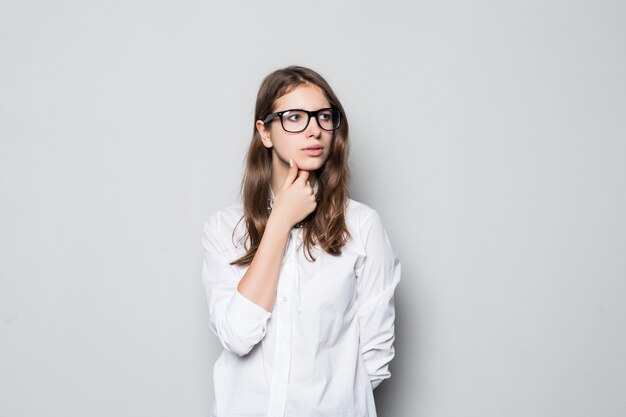 Chica joven con gafas vestidas con una estricta camiseta blanca de oficina se encuentra frente a la pared blanca