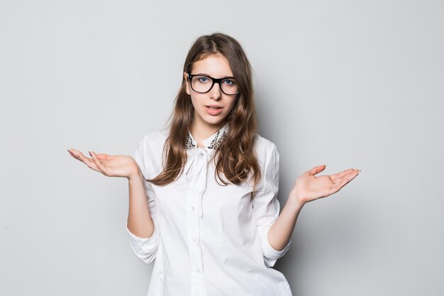 Chica joven con gafas vestidas con una estricta camiseta blanca de oficina se encuentra frente a la pared blanca