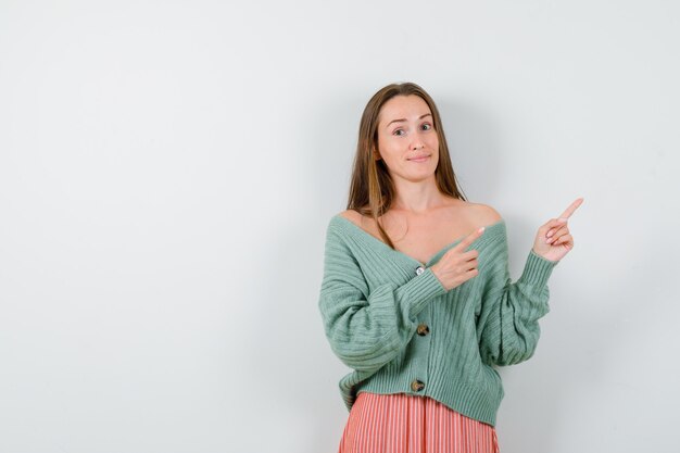 Chica joven en géneros de punto, falda apuntando hacia la derecha con los dedos índices y mirando feliz, vista frontal.