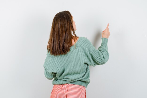 Chica joven en géneros de punto, falda girando hacia atrás, apuntando a la pared con el dedo índice y mirando seductora, vista frontal.