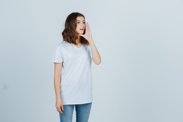 Chica joven gritando a alguien en camiseta blanca, jeans y mirando linda, vista frontal.