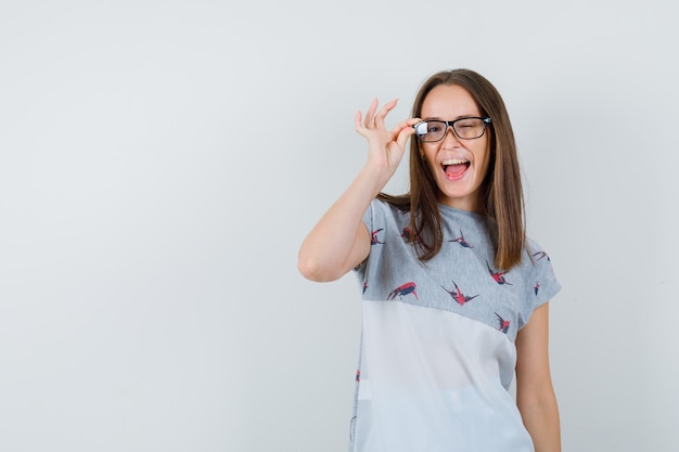 Chica joven guiñando el ojo con los dedos en las gafas en camiseta y mirando loco, vista frontal.