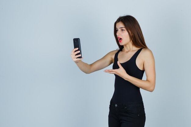 Chica joven hablando con alguien por teléfono, estirando la mano hacia el teléfono en top negro, pantalones y mirando enfocado. vista frontal.