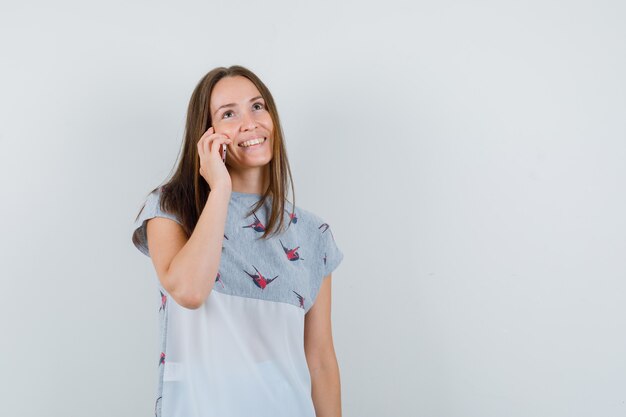 Chica joven hablando por teléfono móvil en camiseta y mirando contento, vista frontal.