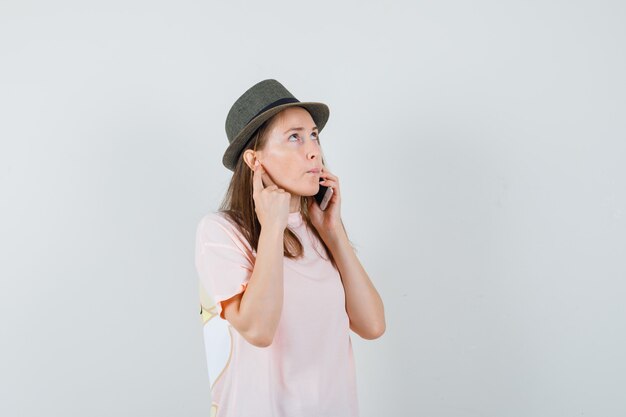 Chica joven hablando por teléfono móvil en camiseta rosa, sombrero y mirando pensativo, vista frontal.