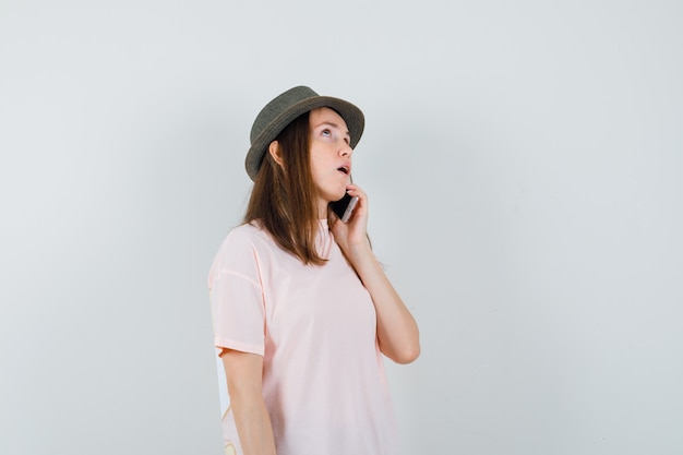 Chica joven hablando por teléfono móvil en camiseta rosa, sombrero y mirando pensativo, vista frontal.