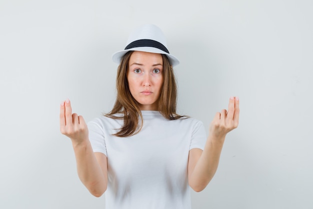 Chica joven haciendo gesto de dinero en camiseta blanca, sombrero y aspecto sensato. vista frontal.