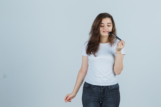 Chica joven haciendo maquillaje con pincel en camiseta, jeans y aspecto encantador, vista frontal.