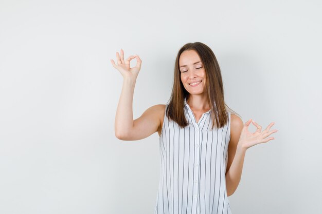 Chica joven haciendo el signo de ok con los ojos cerrados en camiseta y luciendo pacífica. vista frontal.