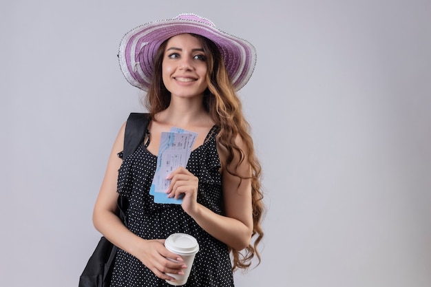 Chica joven hermosa viajera con sombrero de verano con mochila sosteniendo billetes de avión sonriendo alegremente feliz y positivo de pie sobre fondo blanco