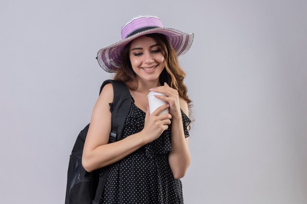 Chica joven hermosa viajera con sombrero de verano con mochila sosteniendo la taza de café sonriendo alegremente feliz y positivo de pie sobre fondo blanco.