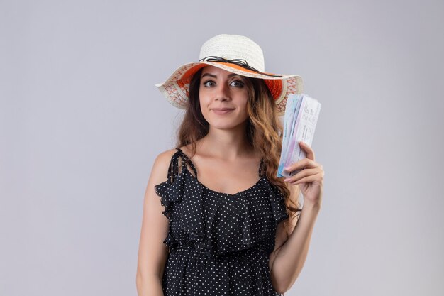 Chica joven hermosa viajera en vestido de lunares en sombrero de verano con billetes de avión mirando a cámara con expresión de confianza en la cara de pie sobre fondo blanco