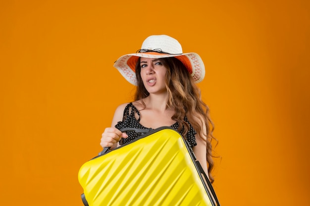 Chica joven hermosa viajera en vestido de lunares en sombrero de verano con maleta mirando a cámara con expresión escéptica de pie sobre fondo amarillo