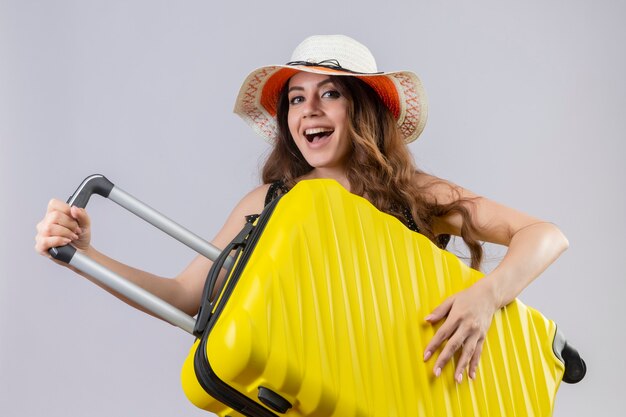 Chica joven hermosa viajera en vestido de lunares en sombrero de verano con maleta mirando a cámara sonriendo alegremente feliz y positivo de pie sobre fondo blanco