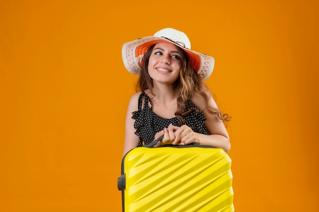 Chica joven hermosa viajera en vestido de lunares en sombrero de verano con maleta mirando a un lado sonriendo alegremente feliz y positivo de pie sobre fondo naranja