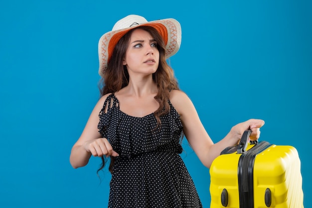 Chica joven hermosa viajera en vestido de lunares en sombrero de verano con maleta nerviosa y muy ansiosa mirando lejos de pie sobre fondo azul