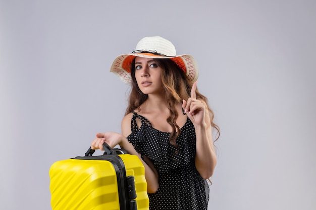 Chica joven hermosa viajera en vestido de lunares en sombrero de verano con maleta de pie con el dedo apuntando hacia arriba concentrado en una tarea de pie sobre fondo blanco