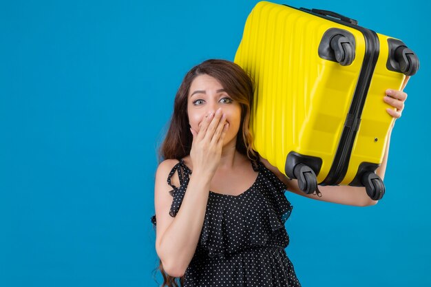 Chica joven hermosa viajera en vestido de lunares sosteniendo maleta mirando sorprendido cubriendo la boca con la mano de pie sobre fondo azul