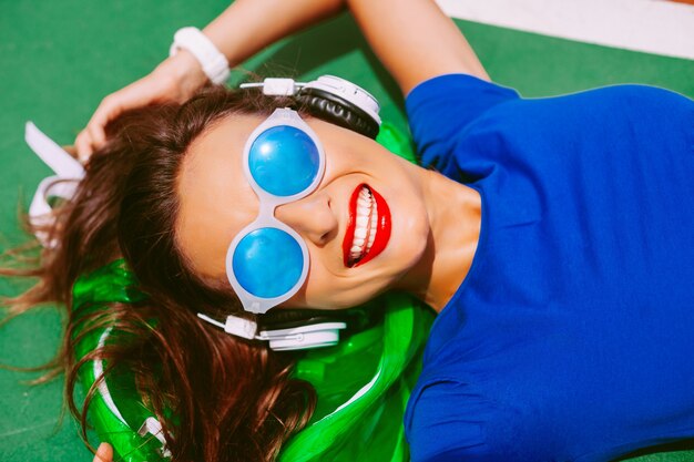 Chica joven inconformista feliz tumbado en su mochila de neón claro, escuchando su música favorita y disfrutando del anuncio de verano de sus vacaciones. Tener dientes blancos perfectos y labios rojos brillantes.