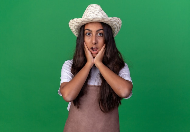 Chica joven jardinero en delantal y sombrero de verano asombrado y sorprendido con las manos en las mejillas de pie sobre la pared verde