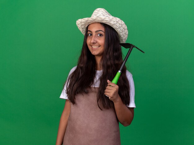 Chica joven jardinero en delantal y sombrero de verano con azadón feliz y alegre sonriente de pie sobre la pared verde