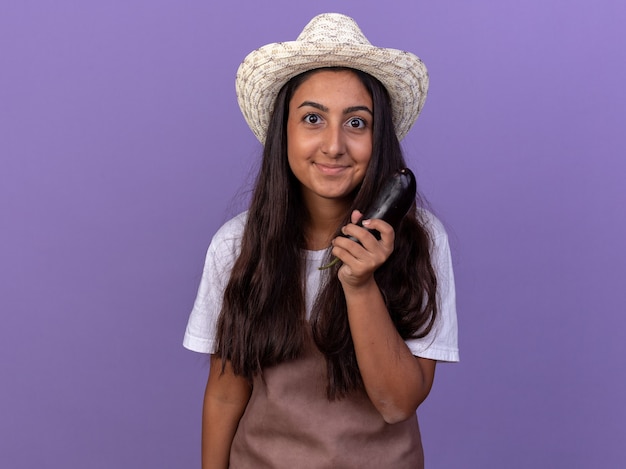 Chica joven jardinero en delantal y sombrero de verano con berenjena feliz y positiva sonriendo de pie sobre la pared púrpura