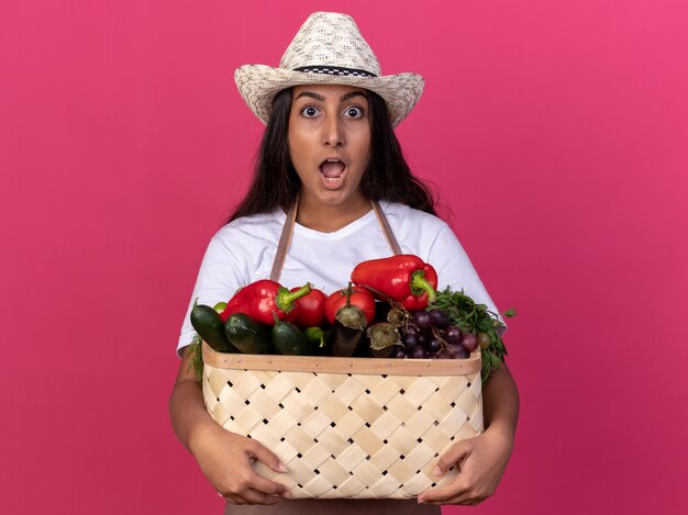 Chica joven jardinero en delantal y sombrero de verano con caja llena de verduras sorprendido y asombrado de pie sobre la pared rosa