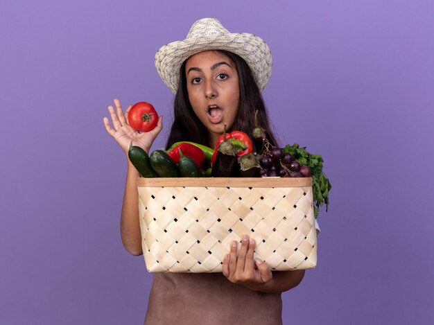 Chica joven jardinero en delantal y sombrero de verano con caja llena de verduras y tomate fresco feliz y sorprendido de pie sobre la pared púrpura