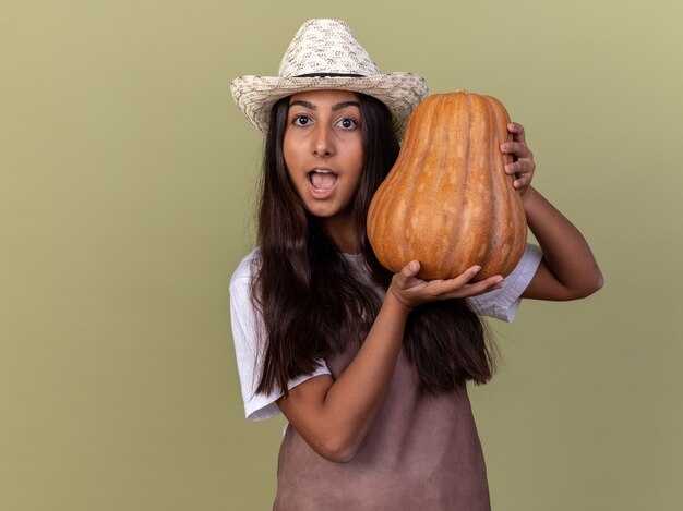 Chica joven jardinero en delantal y sombrero de verano con calabaza de pie asombrado y sorprendido sobre la pared verde