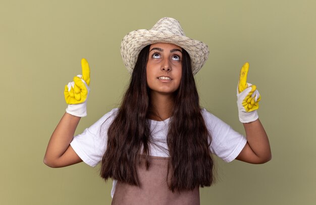 Chica joven jardinero en delantal y sombrero de verano con guantes de trabajo apuntando hacia arriba con los dedos índices mirando hacia arriba con una sonrisa en la cara de pie sobre la pared verde