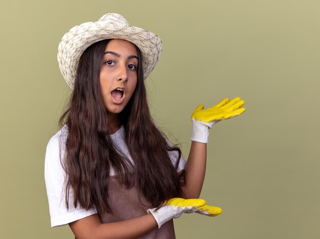 Chica joven jardinero en delantal y sombrero de verano con guantes de trabajo sorprendió presentando algo con los brazos de pie sobre la pared verde