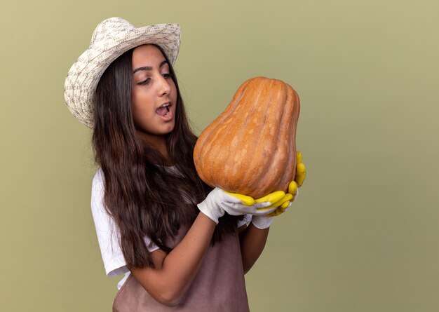 Chica joven jardinero en delantal y sombrero de verano con guantes de trabajo sosteniendo calabaza mirándola asombrada y feliz de pie sobre la pared verde