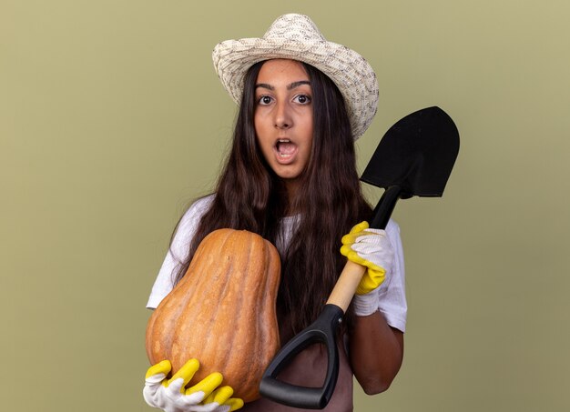 Chica joven jardinero en delantal y sombrero de verano con guantes de trabajo sosteniendo calabaza y pala asombrado y sorprendido de pie sobre la pared verde