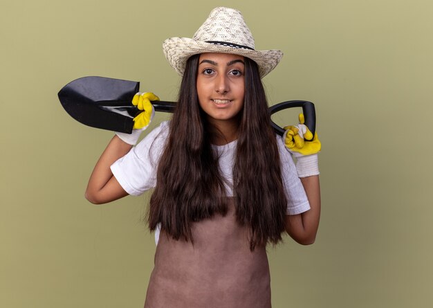 Chica joven jardinero en delantal y sombrero de verano con guantes de trabajo sosteniendo la pala sonriendo confiado de pie sobre la pared verde